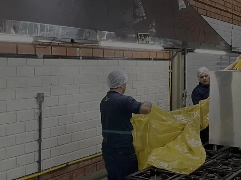 Técnicos da Ecoinset, de touca e uniforme azul-marinho, protegem os equipamentos de uma cozinha industrial antes de limpar a coifa.